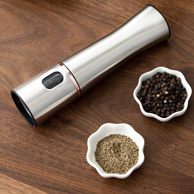 A modern silver electric salt and pepper mill is displayed next to two small white bowls filled with black peppercorns and ground pepper, set on a wooden surface.