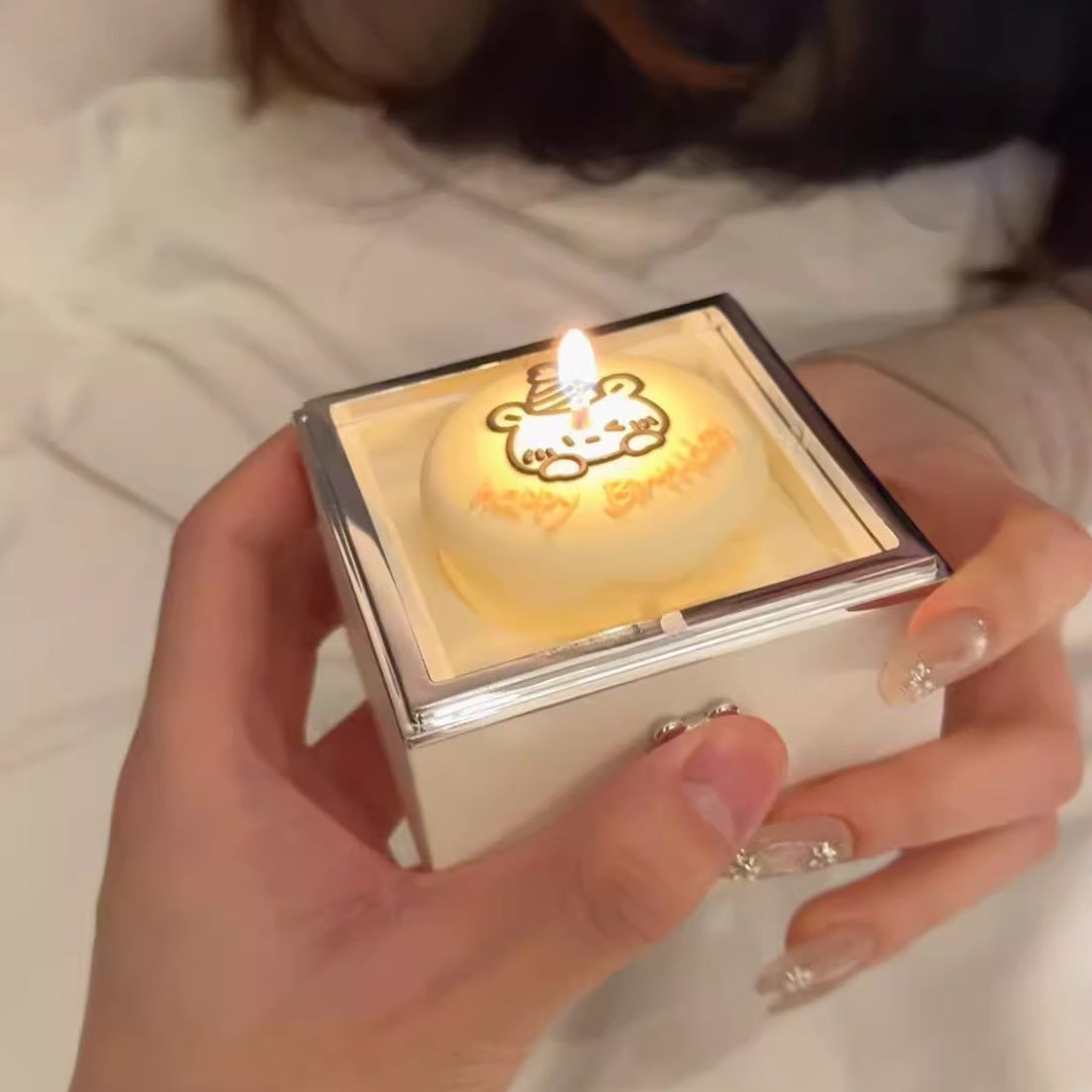 Hand holding a small lit candle in a decorative box with a bear design.