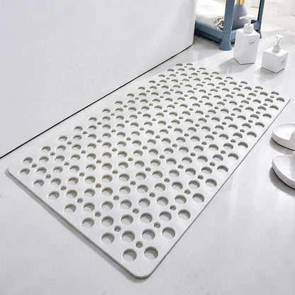 A white rectangular bath mat with a grid of circular drainage holes, shown on a light gray bathroom floor next to white bath accessories.