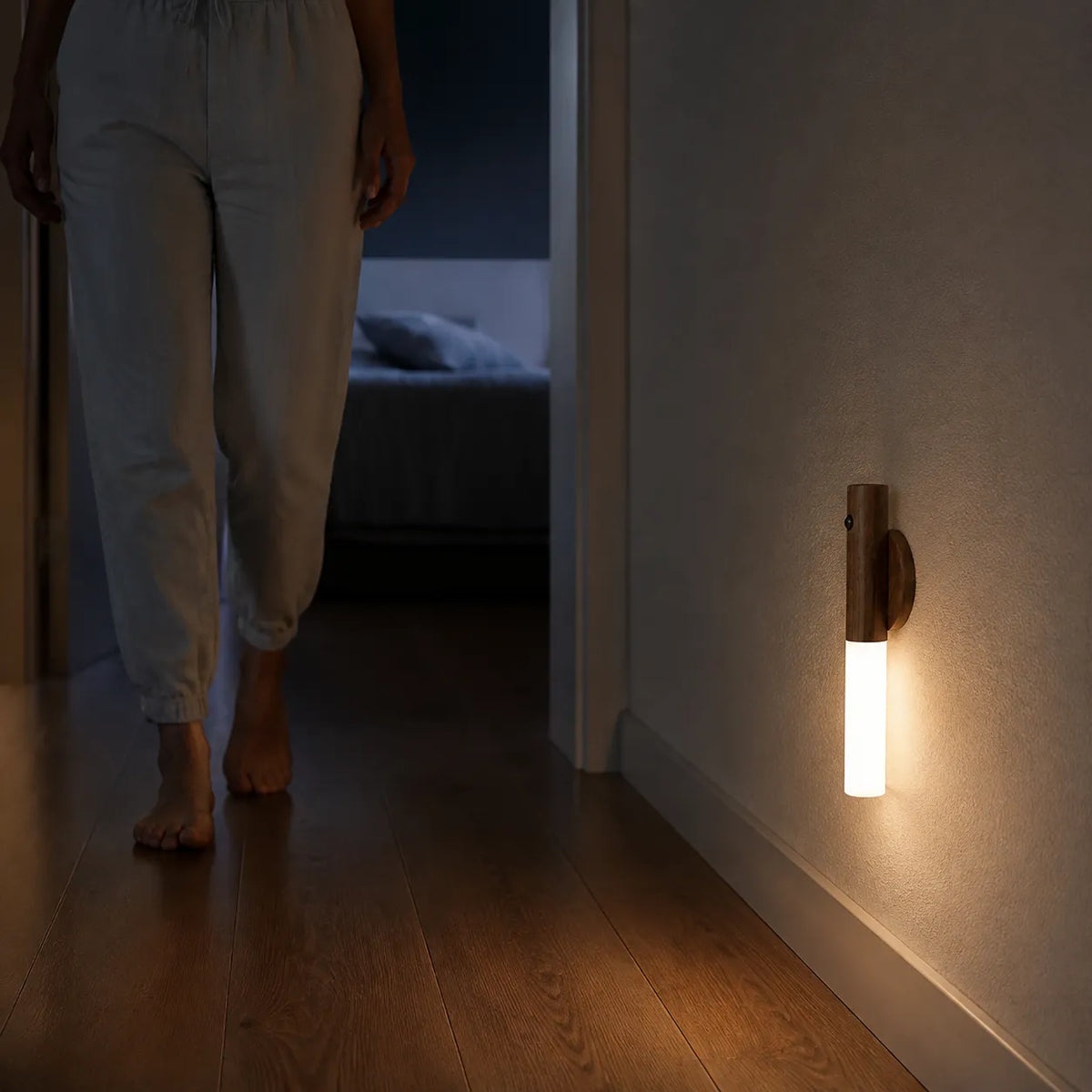 A wall-mounted cylindrical light fixture with a warm glowing vertical tube and a dark wood-toned top, installed on a beige wall in a dimly lit hallway. A person wearing light-colored pants is walking barefoot in the background.