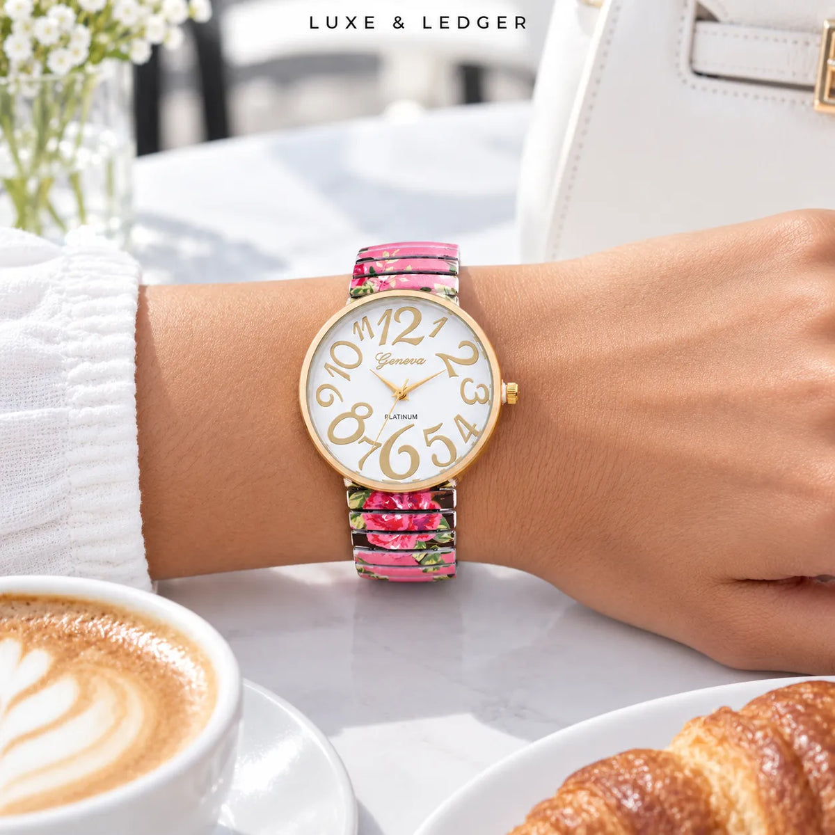A close-up lifestyle shot of a Luxe & Ledger women's watch with a gold-tone case and a pink floral patterned strap, worn on a woman's wrist in a cafe setting.