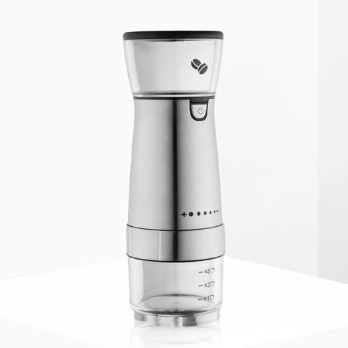 A stainless steel electric coffee grinder with a clear container, displaying the grinding process with coffee beans inside, and a cup and a napkin beside it on a dark background.