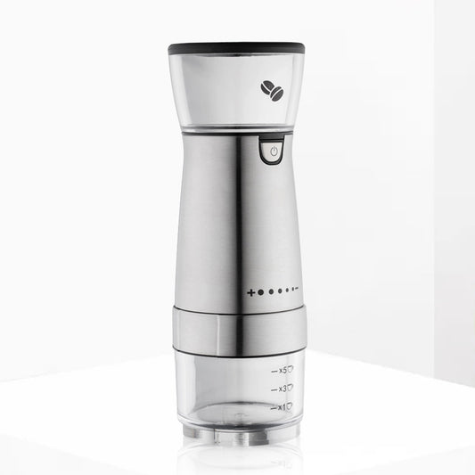 A stainless steel electric coffee grinder with a clear container, displaying the grinding process with coffee beans inside, and a cup and a napkin beside it on a dark background.