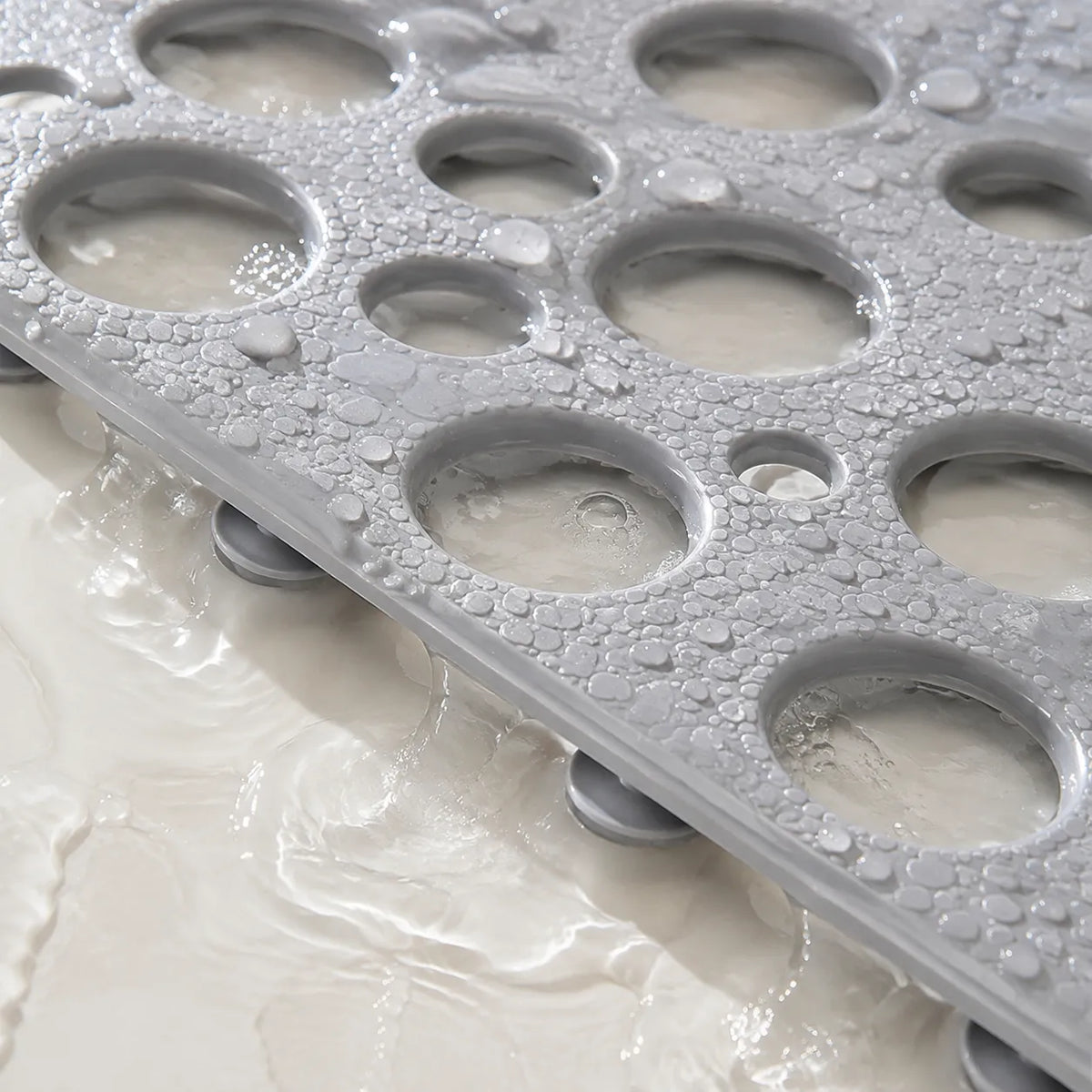 A close-up of a gray silicone bath mat with multiple oval drainage holes, shown with water draining off onto a white surface. The mat has a textured surface covered in water droplets.