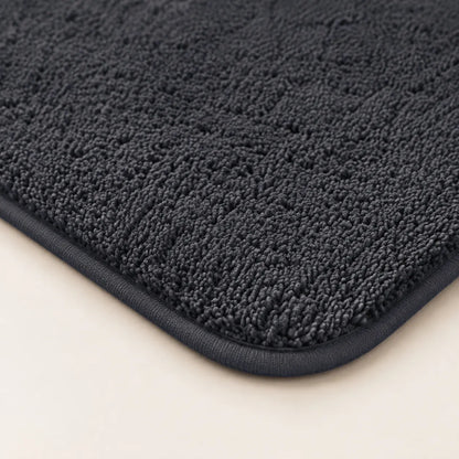 A close-up of a dark charcoal gray textured bath mat corner with a thick dark border, shown on a light beige background.