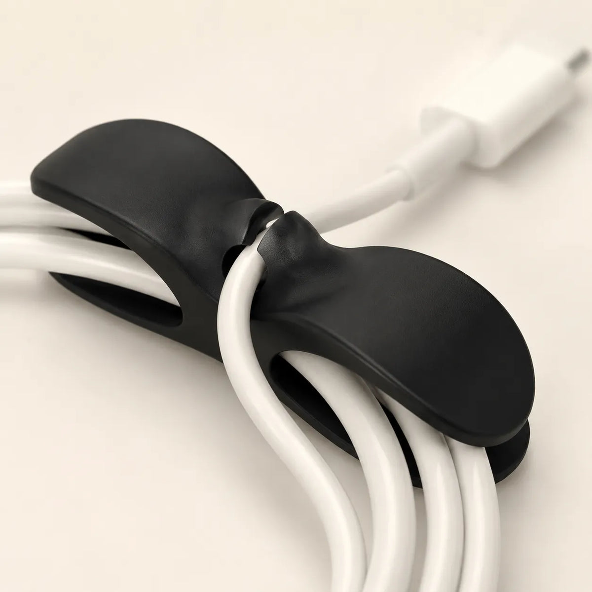 A close-up of a black cable organizer clip holding several white cables, with a white connector visible at the top right on a plain light background.