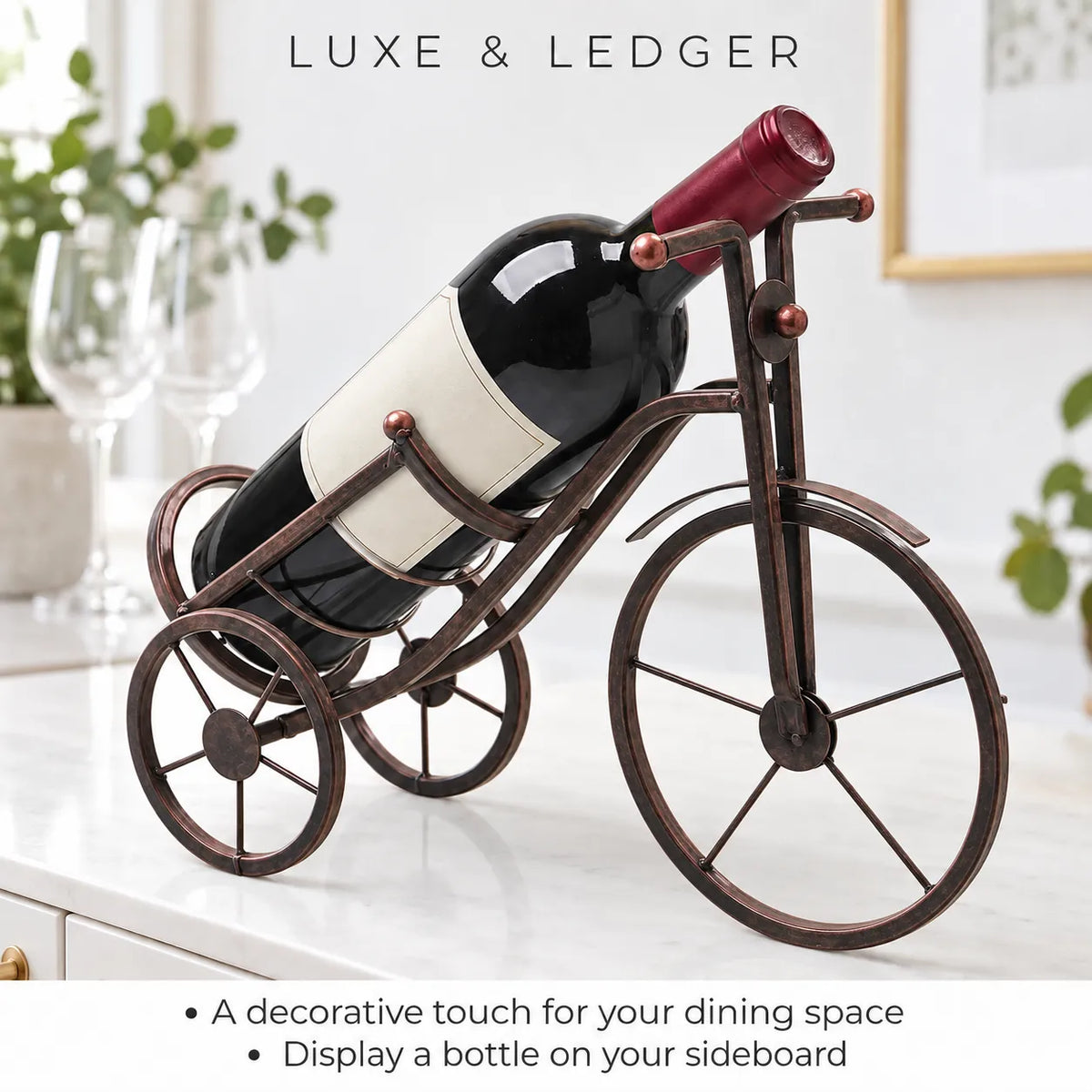 A wine bottle displayed on a decorative metal tricycle stand shaped like a bicycle, set on a white countertop in a bright kitchen or dining space. The stand is dark bronze with copper-toned accents.