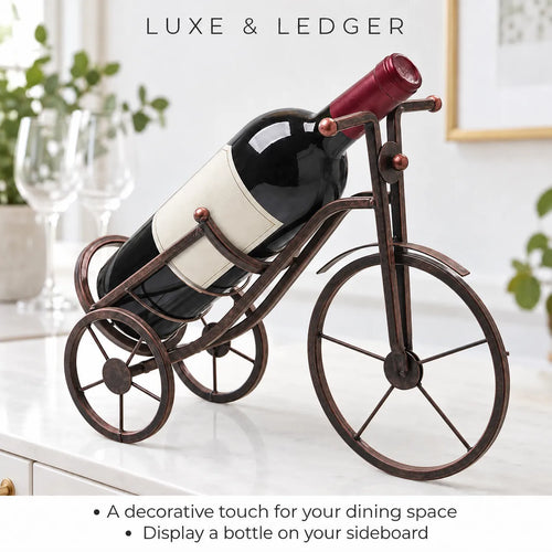 A wine bottle displayed on a decorative metal tricycle stand shaped like a bicycle, set on a white countertop in a bright kitchen or dining space. The stand is dark bronze with copper-toned accents.