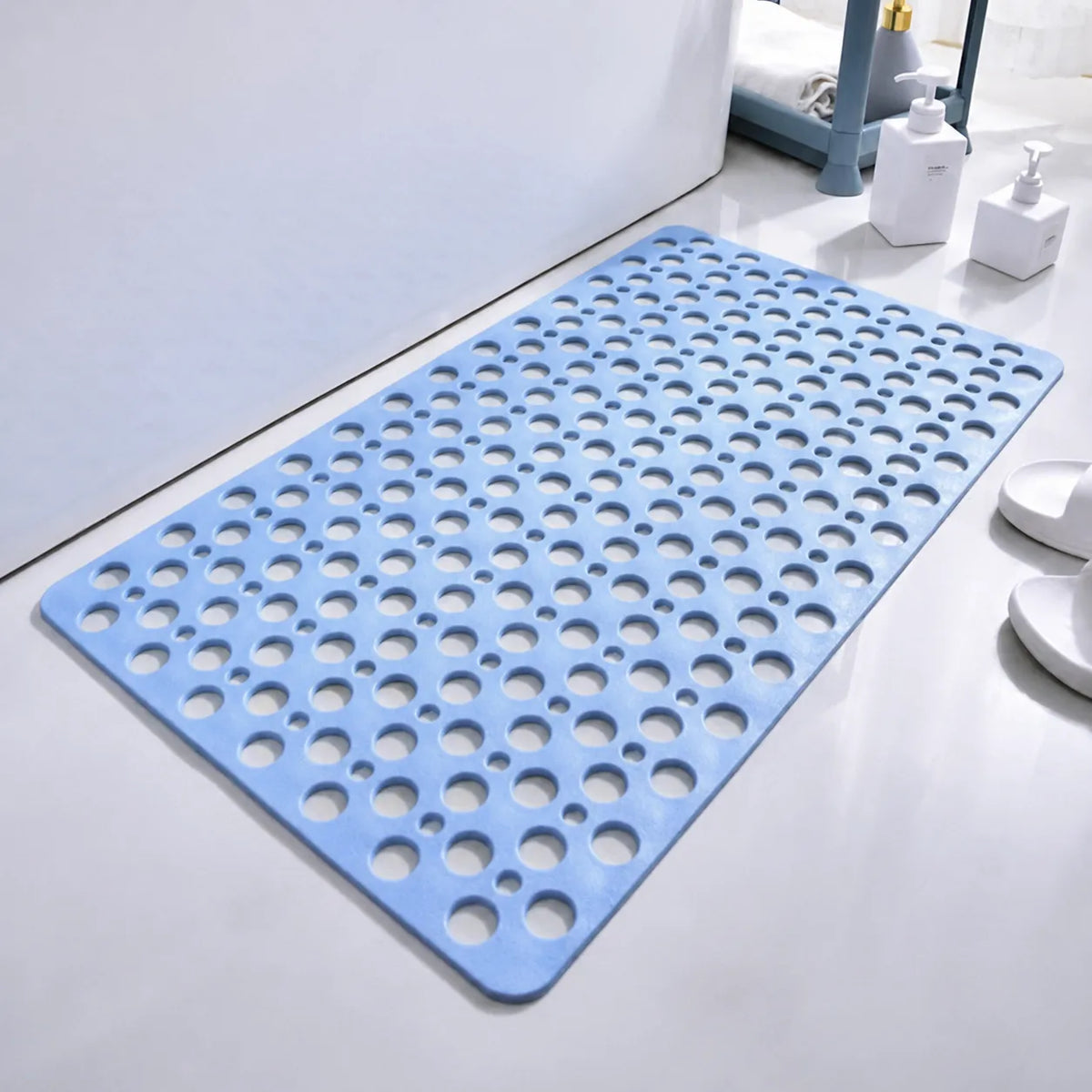 A light blue bath mat with a raised circular drainage pattern, shown on a white bathroom floor next to a bathtub, with white slippers and bathroom accessories in the background.