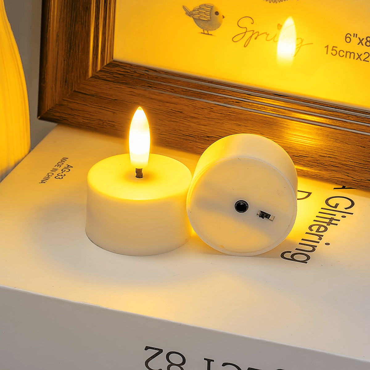 Two small round candles with visible flames on a surface with a wooden frame in the background.