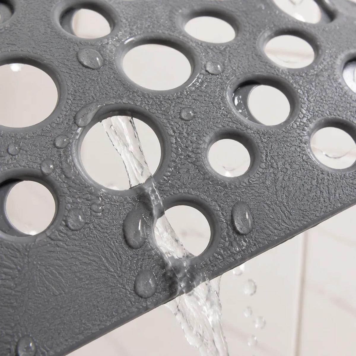 Close-up of a dark gray textured bath mat with circular drainage holes, shown with water pouring through the holes against a white tiled background.