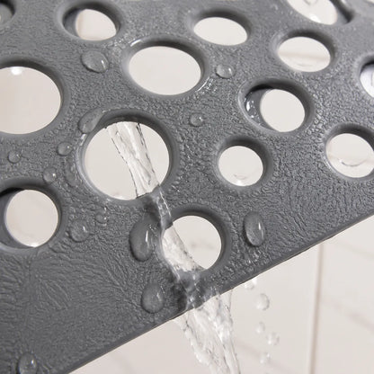 Close-up of a dark gray textured bath mat with circular drainage holes, shown with water pouring through the holes against a white tiled background.