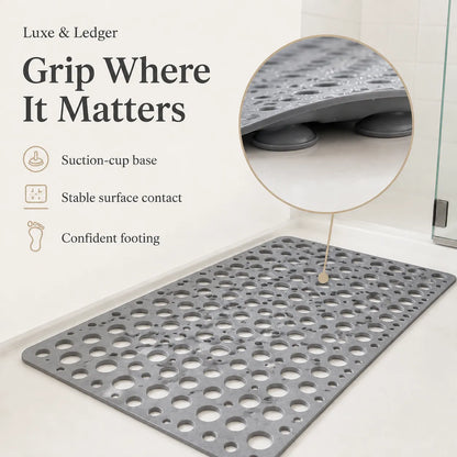 A gray textured bath mat with circular drainage holes shown on a bathroom floor, with a circular inset highlighting the suction-cup base. Text on the left reads 'Grip Where It Matters' and lists features including suction-cup base, stable surface contact, and confident footing.