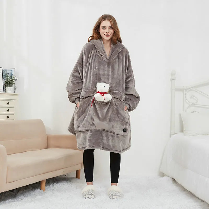 A woman stands in a bright bedroom wearing a plush hooded robe in taupe-gray. The robe has large front pockets, a small white plush bear pocket with a red ribbon, and is paired with black leggings and white slippers.
