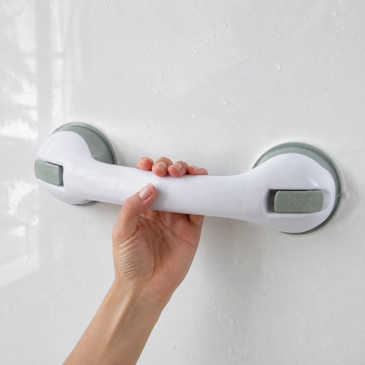 A hand holding a white grab handle with gray suction cups mounted on a white tiled wall with water droplets.