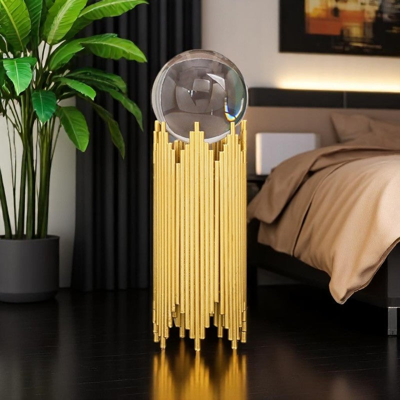Gold decorative lamp on a dark wooden floor with a plant and bed in the background