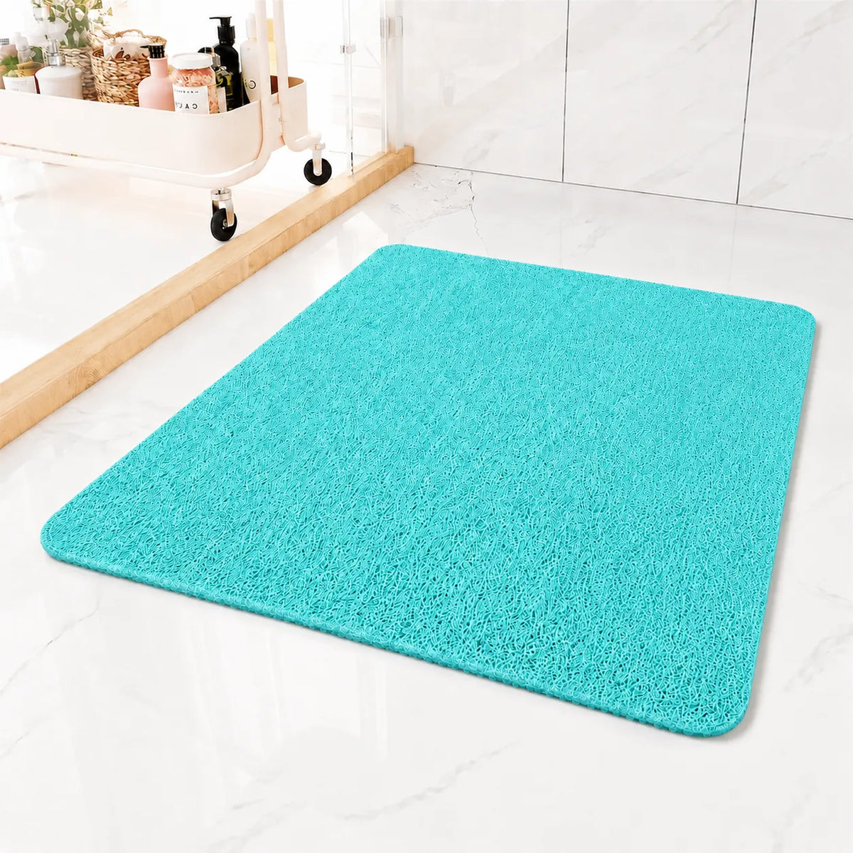 A bright turquoise blue rectangular bath mat with rounded corners, shown on a white marble-look floor in a bathroom setting next to a wooden-framed mirror and toiletry cart.
