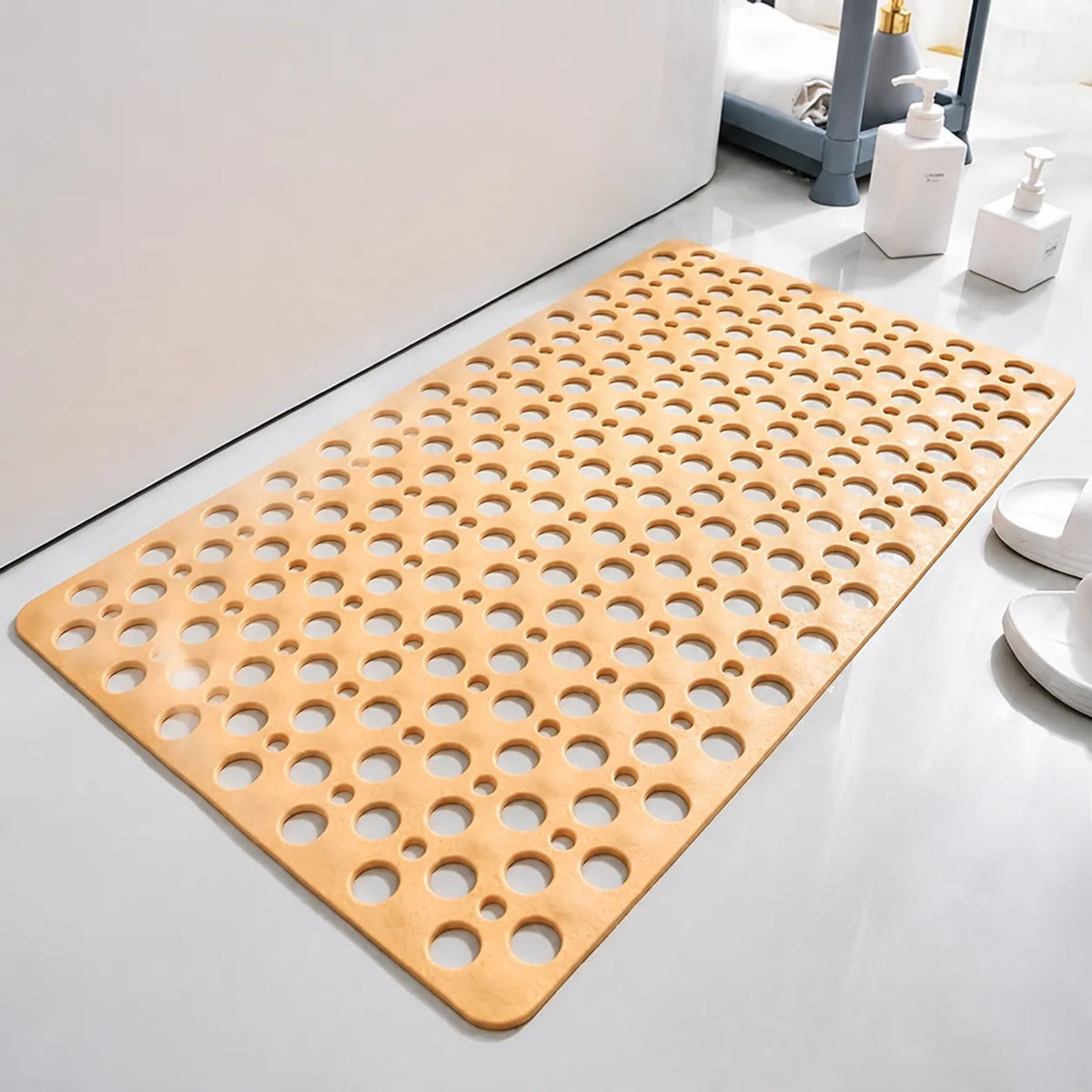 A rectangular bath mat with a light natural wood finish and a grid of circular cutouts, shown on a bathroom floor next to a bathtub, toiletry bottles, and white slippers.
