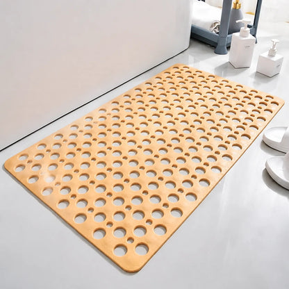 A rectangular bath mat with a light natural wood finish and a grid of circular cutouts, shown on a bathroom floor next to a bathtub, toiletry bottles, and white slippers.
