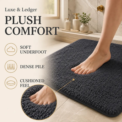 A plush bath mat in dark charcoal gray shown in a bathroom setting with a person's foot stepping onto it. The image includes marketing text reading 'PLUSH COMFORT' and feature callouts for soft underfoot, dense pile, and cushioned feel, plus a circular close-up of the mat texture.