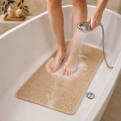 A beige bath mat inside a white bathtub while a person showers, with water running over the mat. The person's legs and hands are visible, and the scene is set in a real bathroom environment.
