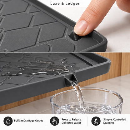 A close-up of a dark gray silicone kitchen draining mat with a finger pressing a release button, shown in two panels. The top panel includes the text 'Luxe & Ledger' and the bottom panel shows water draining into a glass, with feature icons for 'Built-in Drainage Outlet', 'Press to Release Collected Water', and 'Simple, Controlled Draining'.