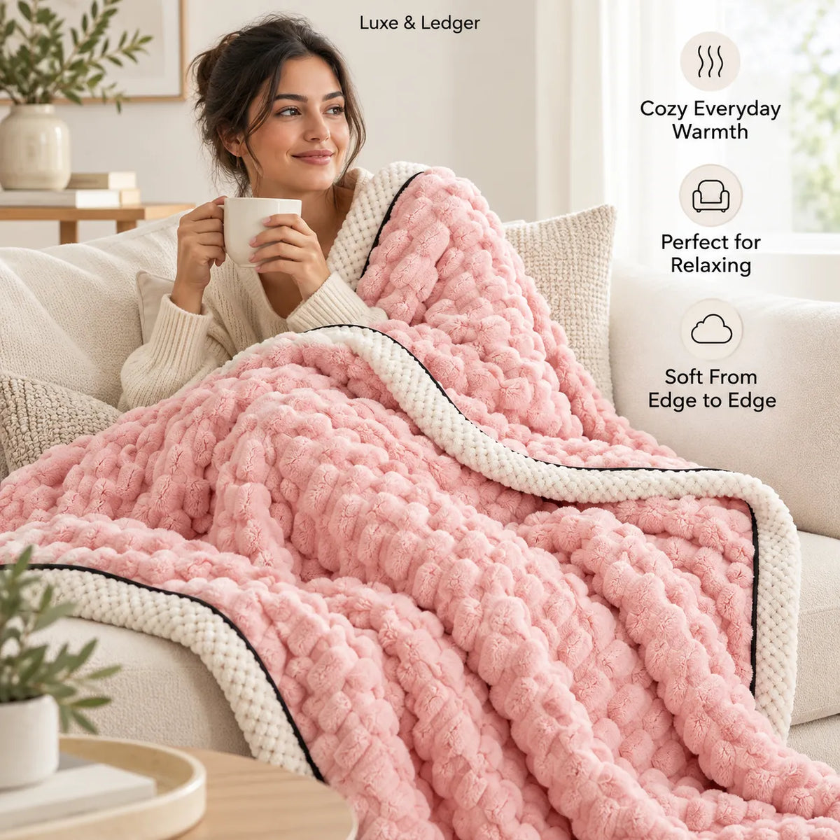 A woman sits on a cream sofa wrapped in a chunky pink textured blanket with cream and black trim, holding a white mug. Text callouts on the right describe cozy warmth, relaxing use, and soft edge-to-edge coverage.