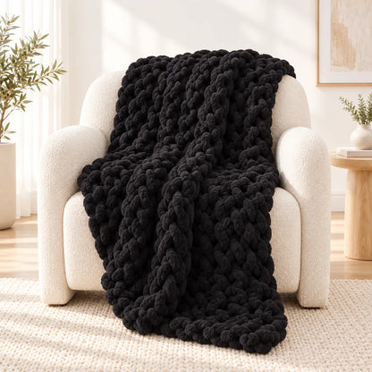 A chunky black knit throw blanket draped over a cream boucle armchair in a bright, modern living room setting.