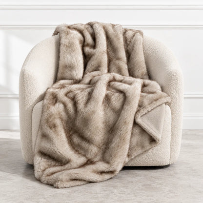 A plush faux fur throw blanket in a warm taupe-beige color, draped over a cream-colored armchair in a bright, modern room.