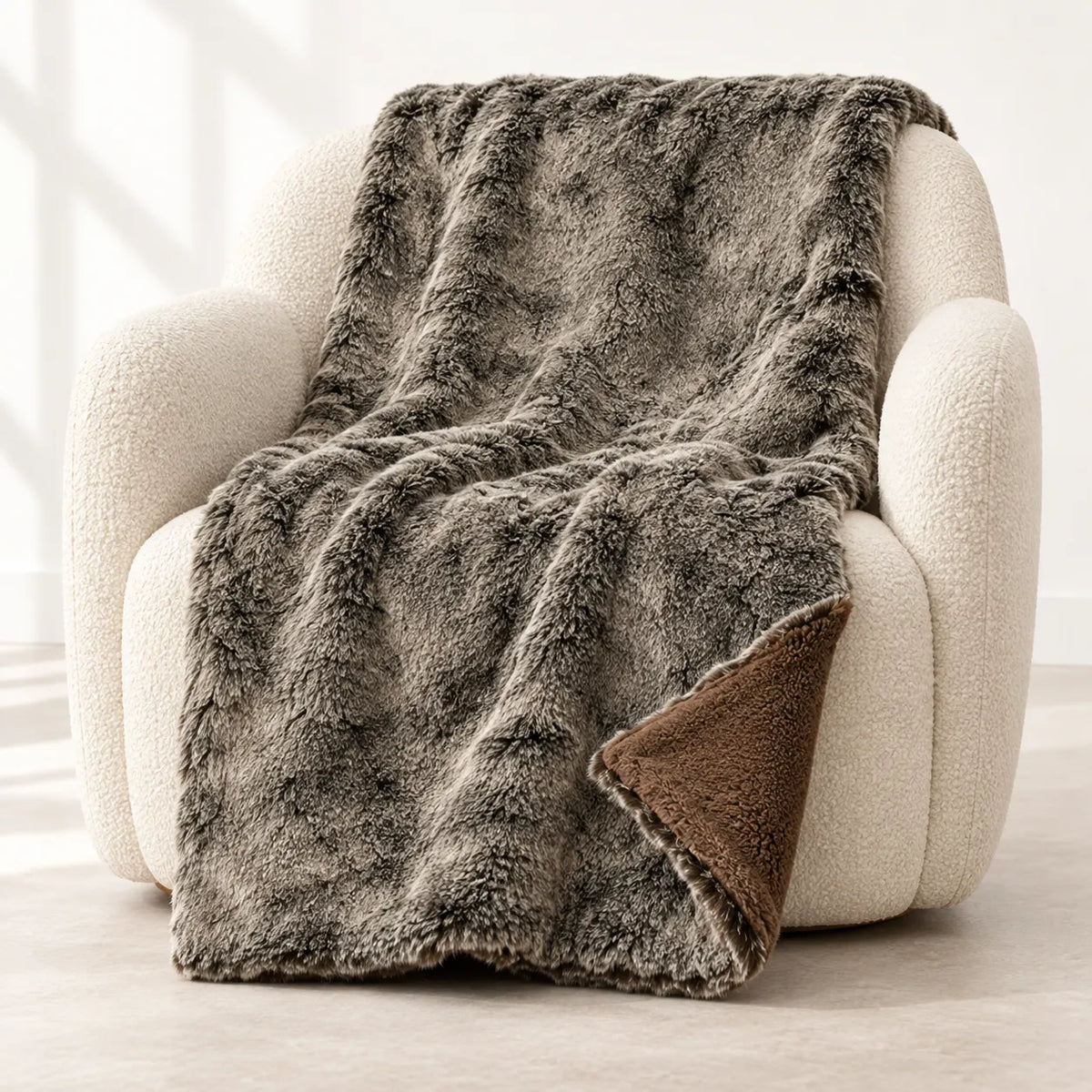 A plush faux-fur throw blanket in a heathered gray-brown color is draped over a cream boucle armchair, showing a brown sherpa lining on the underside.