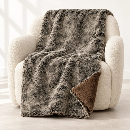 A plush faux-fur throw blanket in a heathered gray-brown color is draped over a cream boucle armchair, showing a brown sherpa lining on the underside.