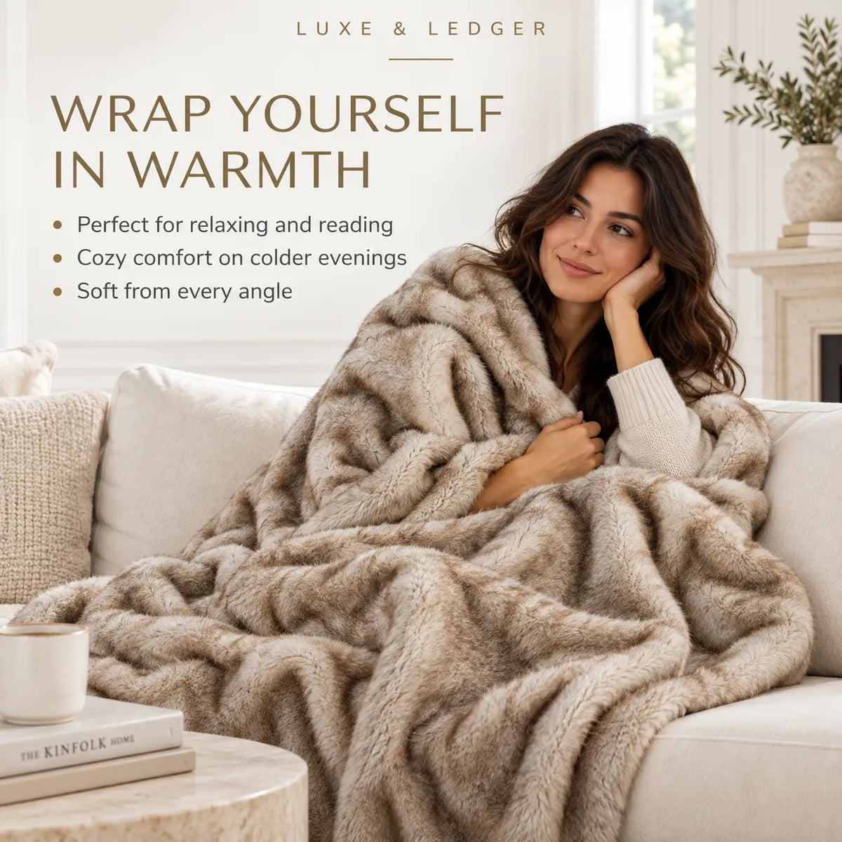 A woman sits on a light sofa wrapped in a plush, textured beige throw blanket. The scene is styled as a cozy living room with a coffee table, mug, and fireplace in the background.