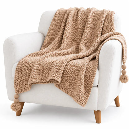 A chunky knit throw blanket in a warm camel beige color, draped over a white armchair with light wood legs, set against a plain white background.