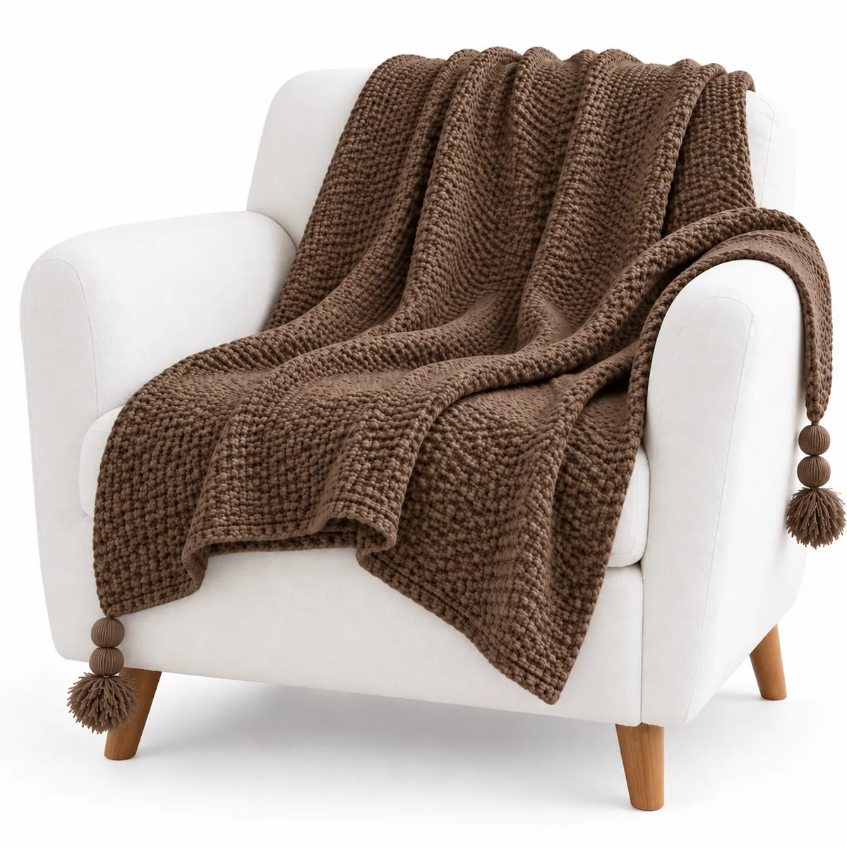 A chunky knit brown throw blanket with pom-pom tassels draped over a white armchair with light wood legs, photographed on a clean white background.
