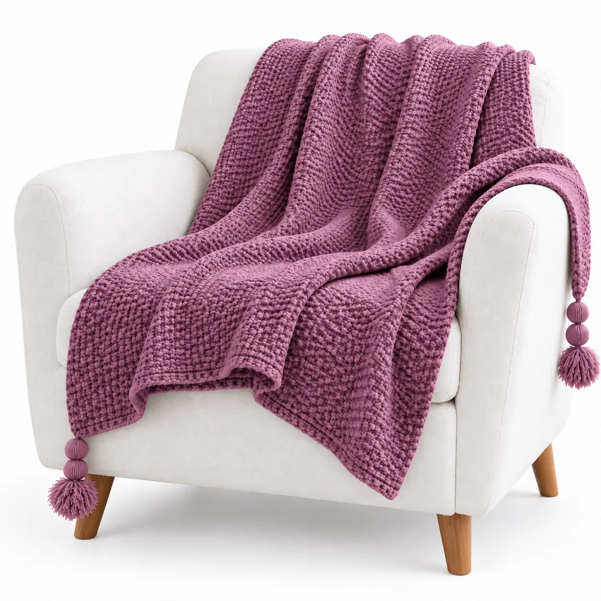 A chunky knit throw blanket in dusty mauve is draped over a white armchair with wooden legs against a clean white background.