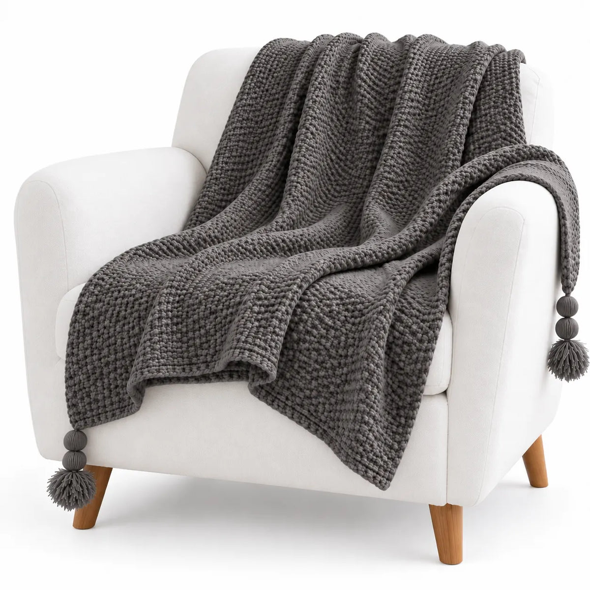 A chunky knit throw blanket in dark gray with pom-pom tassels, draped over a white armchair with wooden legs against a clean white background.