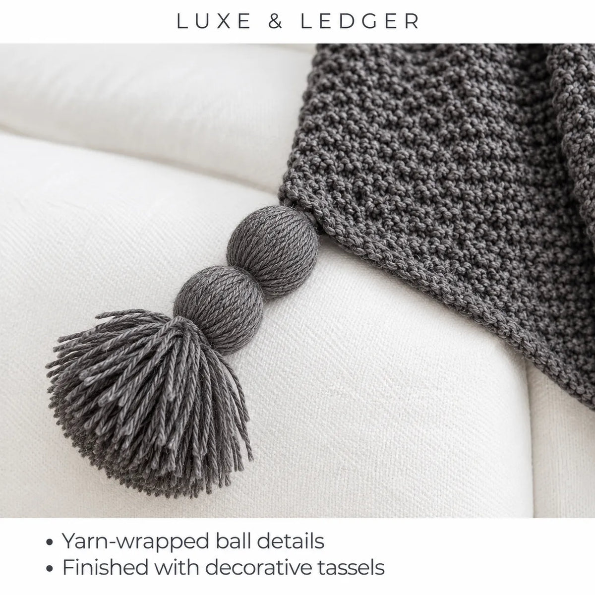 A close-up of a dark gray knitted throw blanket draped over a white upholstered chair, with a large gray tassel detail hanging from the corner. Text at the top reads 'LUXE & LEDGER' and bullet points at the bottom mention yarn-wrapped ball details and decorative tassels.