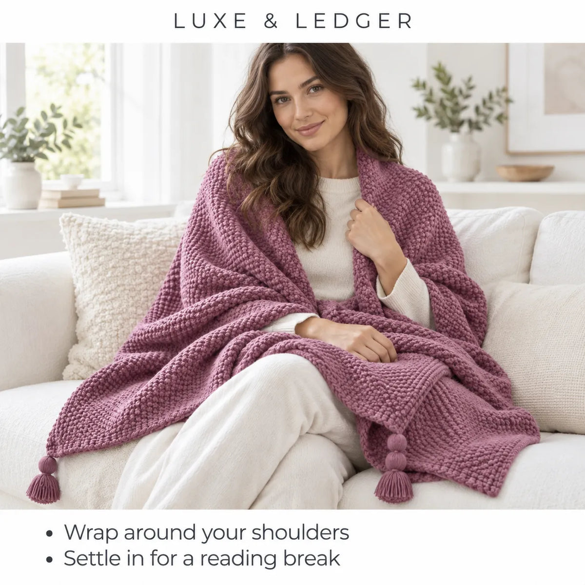 A woman sits on a white sofa wrapped in a chunky knit throw blanket. The blanket is a dusty mauve-purple color with matching tassels at the corners.