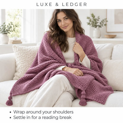A woman sits on a white sofa wrapped in a chunky knit throw blanket. The blanket is a dusty mauve-purple color with matching tassels at the corners.
