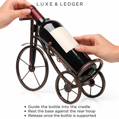 A metal wine bottle carrier shaped like a bicycle, shown with hands guiding a wine bottle into its cradle. The product is displayed on a clean white background with instructional text overlays at the top and bottom.