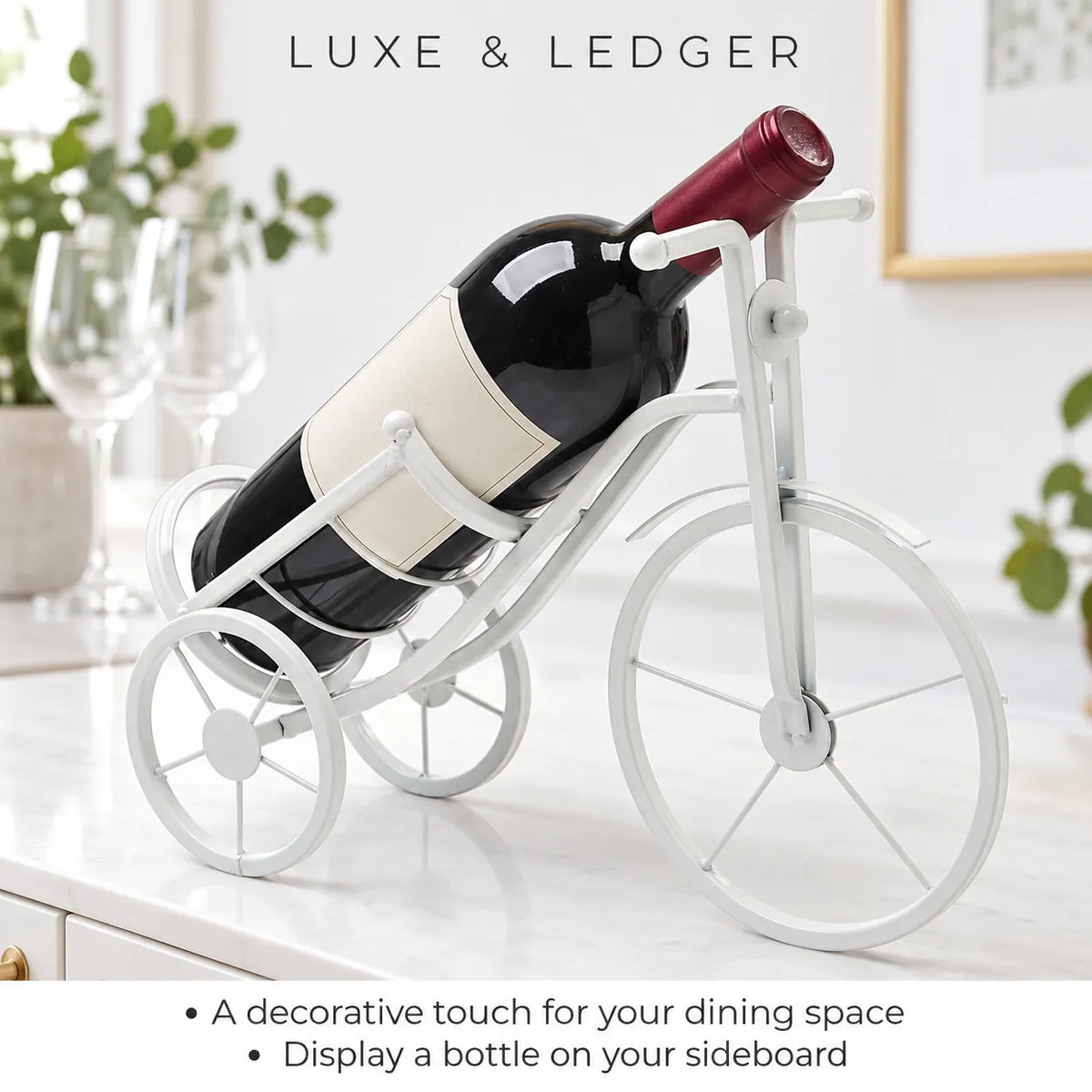 A white bicycle-shaped wine bottle holder displayed on a white sideboard, holding a wine bottle. The background shows a softly blurred kitchen setting with wine glasses and green plants.