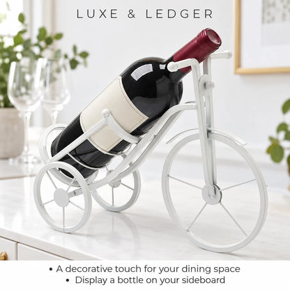 A white bicycle-shaped wine bottle holder displayed on a white sideboard, holding a wine bottle. The background shows a softly blurred kitchen setting with wine glasses and green plants.