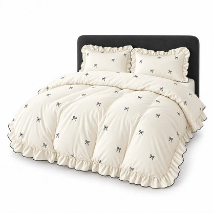 An off-white bedding set with a ruffled edge and small black bow pattern, shown on a bed with a black headboard against a white background.