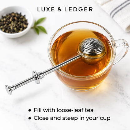 A stainless steel tea infuser with a long handle and spherical mesh ball rests inside a clear glass cup filled with amber tea on a light marble surface. A small bowl of loose-leaf tea and green leaves appear blurred in the background, with the text 'LUXE & LEDGER' at the top and usage instructions at the bottom.