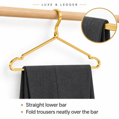 Gold-tone metal trouser hanger hanging from a wooden rod, with folded dark gray trousers draped over the bar. The image includes a circular inset showing a close-up of the hanger fabric and text reading 'LUXE & LEDGER' plus feature bullets.