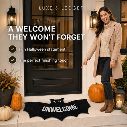 A woman stands at a front door wearing a beige coat and jeans, pointing to a black bat-shaped doormat with the word 'UNWELCOME' in white. Pumpkins, lanterns, and fall decor decorate the porch, with promotional text on the left reading 'A WELCOME THEY WON'T FORGET' and 'LUXE & LEDGER' at the top.