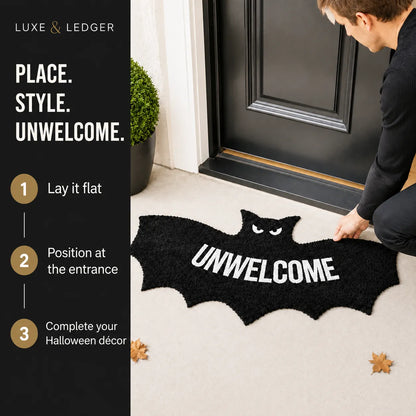 A black bat-shaped doormat with the word "UNWELCOME" in white is being placed at a front door by a person. The left side includes a black panel with the brand name Luxe & Ledger and step-by-step placement instructions.