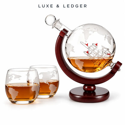 A luxury whiskey decanter and two matching glasses on a white background. The decanter features a clear globe-shaped glass body with an etched world map design, a miniature ship model inside, and amber liquid.
