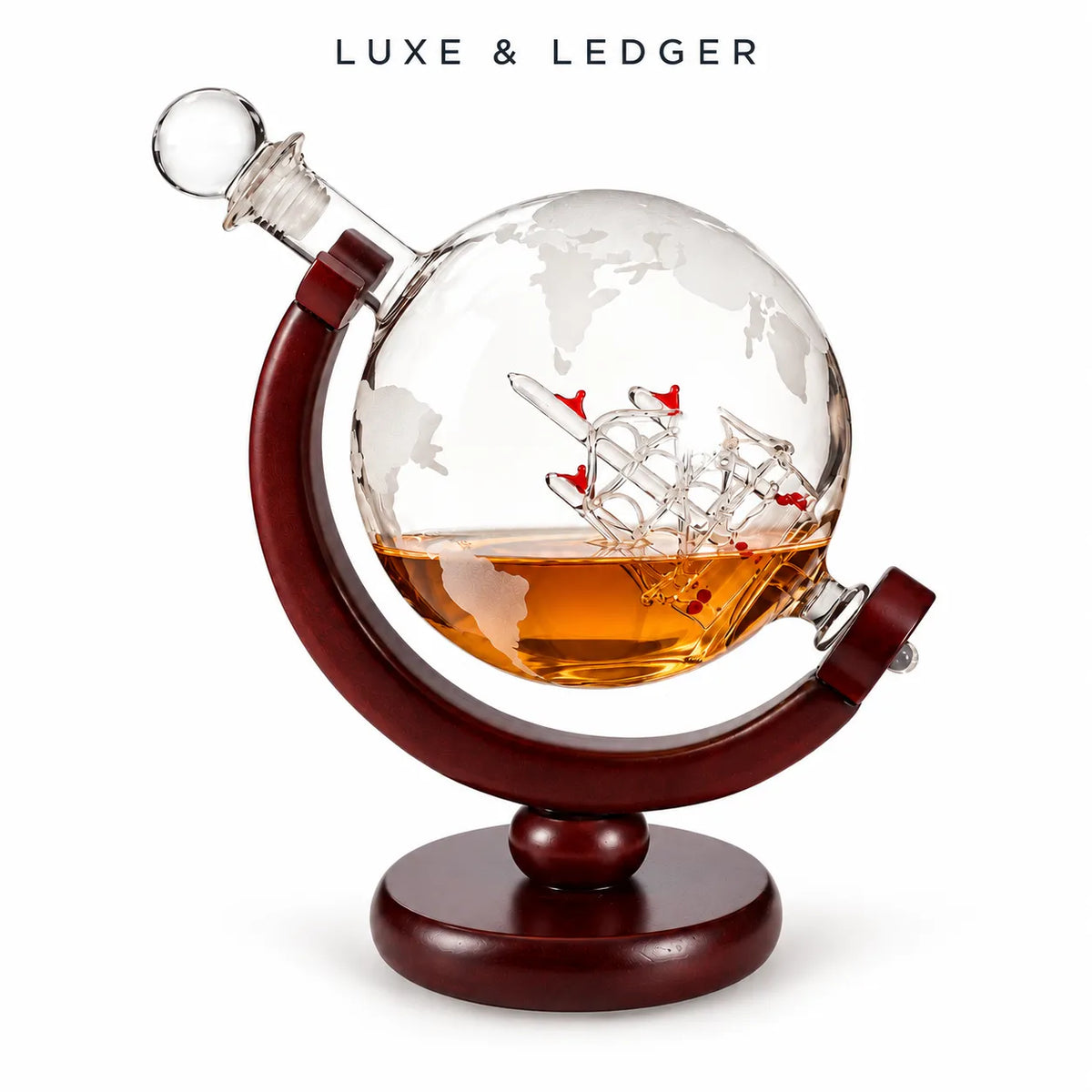 A Luxe & Ledger decorative globe-shaped glass decanter with a world map design, filled with amber liquid and featuring a miniature ship model inside, mounted on a dark reddish-brown wooden stand against a white background.