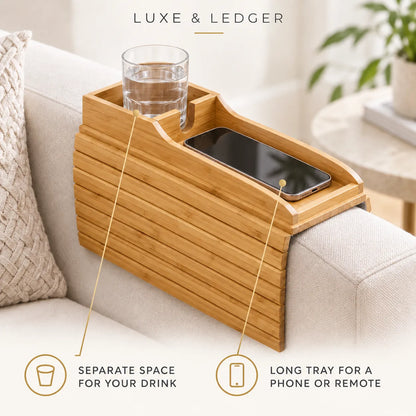 A bamboo armrest tray with a built-in cup holder and phone slot, shown resting on a light beige sofa. The image includes text overlays reading 'LUXE & LEDGER' and feature callouts: 'SEPARATE SPACE FOR YOUR DRINK' and 'LONG TRAY FOR A PHONE OR REMOTE'.